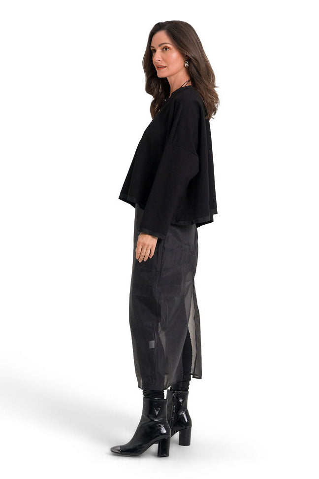 A woman modeling a black long-sleeve draped top, dark gray leather skirt, and black heeled boots against a solid white background.