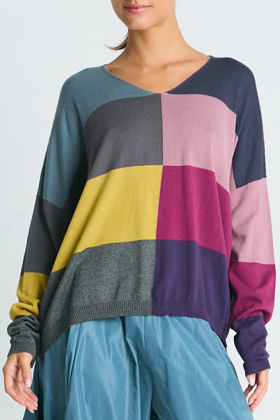 A model wearing a multicolored patchwork V-neck sweater with long sleeves, featuring a color block design with multiple colors including yellow, pink, blue, and gray.