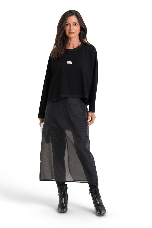 A woman modeling a black long-sleeve scuba top with a small white logo on the chest, paired with a sheer black midi skirt and black ankle boots, against a plain white studio background.