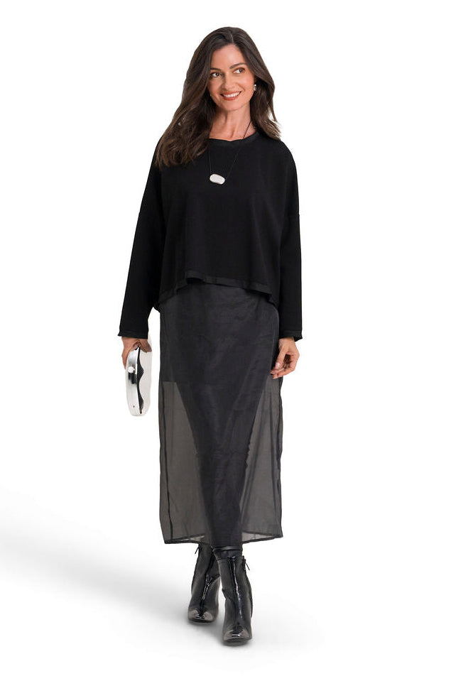 A woman modeling a black long-sleeve top and a sheer black midi skirt, holding a small white clutch, against a solid white background.