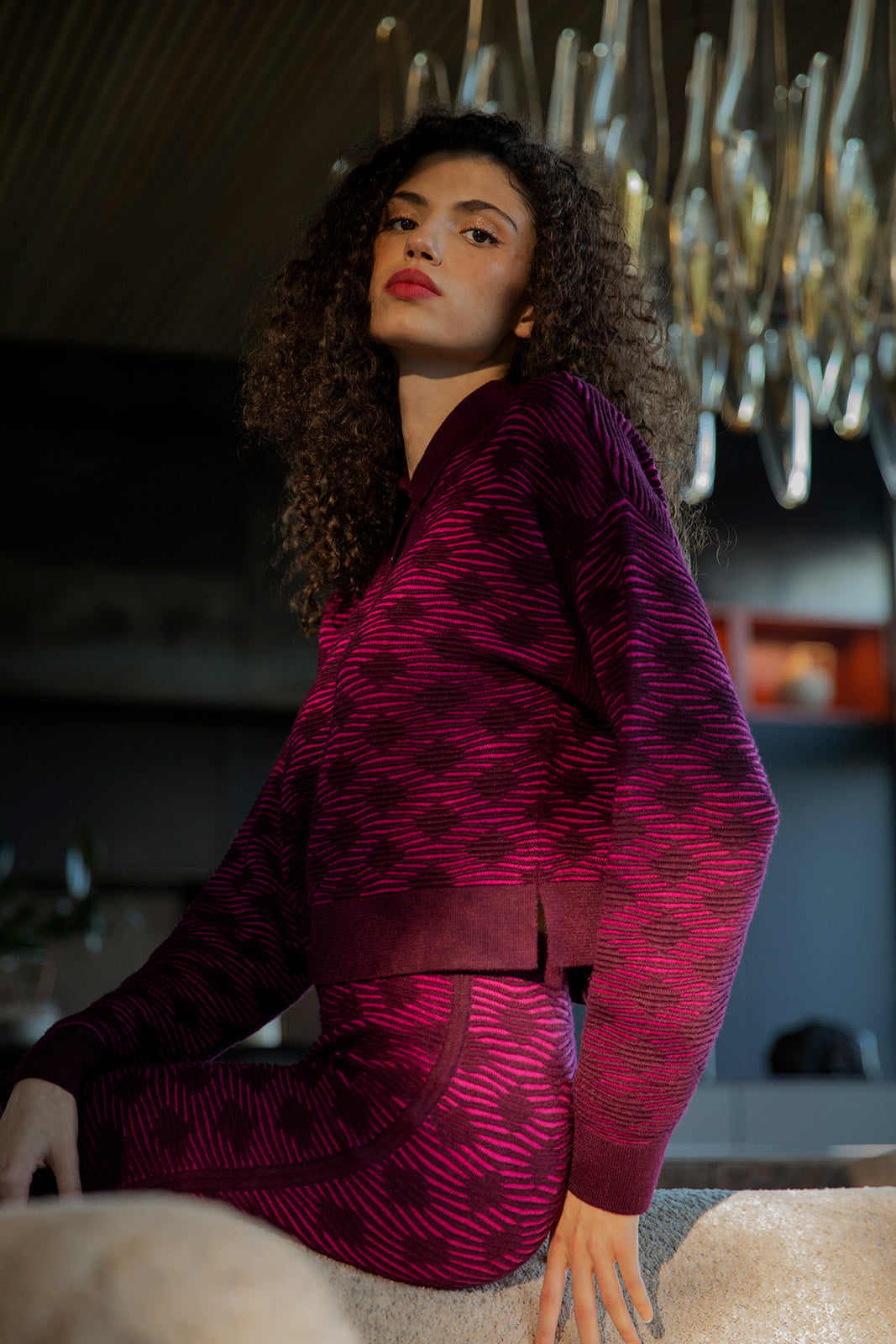 Model sitting on the edge of a beige couch, wearing a matching fuchsia aubergine long sleeve polo sweater and pencil skirt.