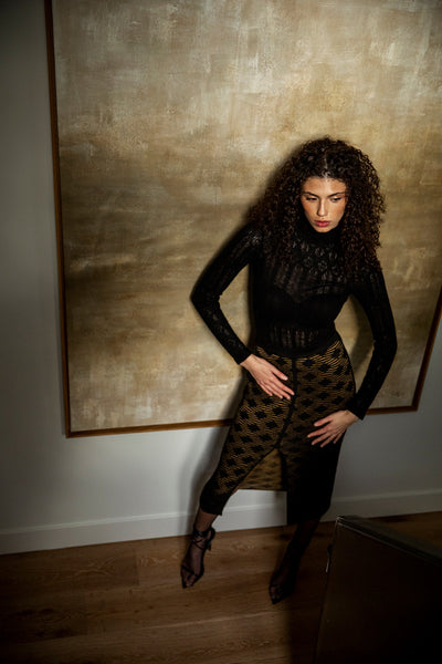 Model in front of a large gold painting, wearing a black lace pointelle pullover top with a mock neck, made from cashmere, styled with a high-waisted pencil skirt in golden black.