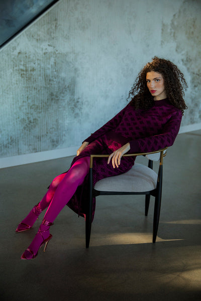 Curly-haired brunette model sitting on a chair wearing a matching fuchsia aubergine set, the Ines Skirt and Amelie Polo both ethically made from baby alpaca and cotton.