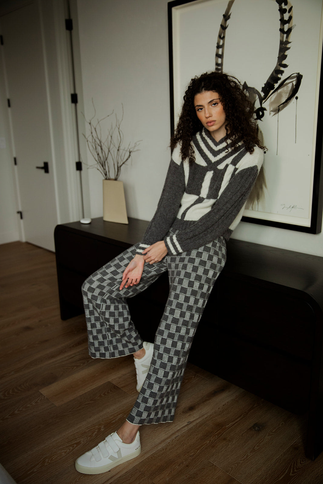 Brunette model sitting indoors against a credenza, wearing the Kari Plaid V Neck Pullover in grey ivory, paired with matching grey reversible trousers and white Veja sneakers.