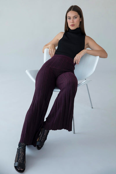 Model sitting on a white chair, styled in aubergine trousers and a black turtleneck bodysuit.