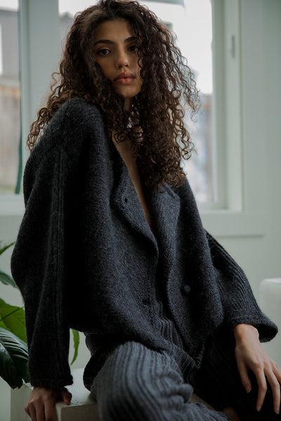 Model Bella sitting down wearing the Momo Handknit in Charcoal. The relaxed, cropped fit and hand-crocheted buttons highlight the artisanal craftsmanship. The cardigan can also be worn upside down for a draped neck.