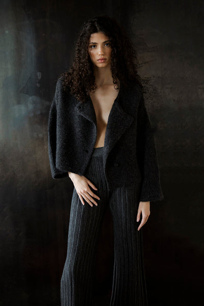 Model standing against a black wall, wearing the Momo Handknit Cardigan in Charcoal open over matching grey trousers known as the Marcelle Trousers by Sskein. Sustainably made in Peru.