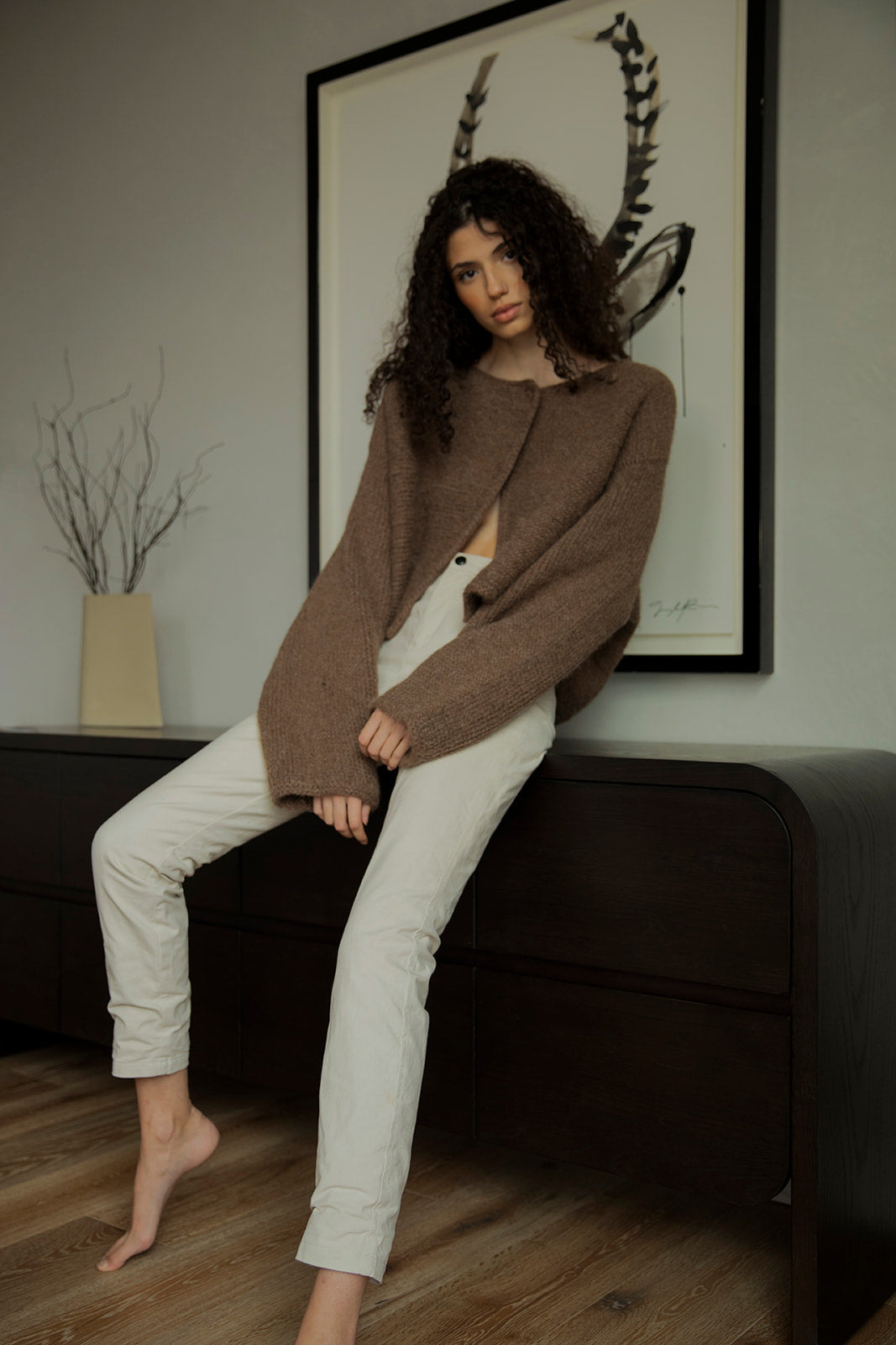 Model wearing the Momo Handknit Cardigan in Saddle, paired with white jeans, standing in a bedroom with a painting in the background. Highlighting an effortless look, the cardigan can also be worn upside down. 
