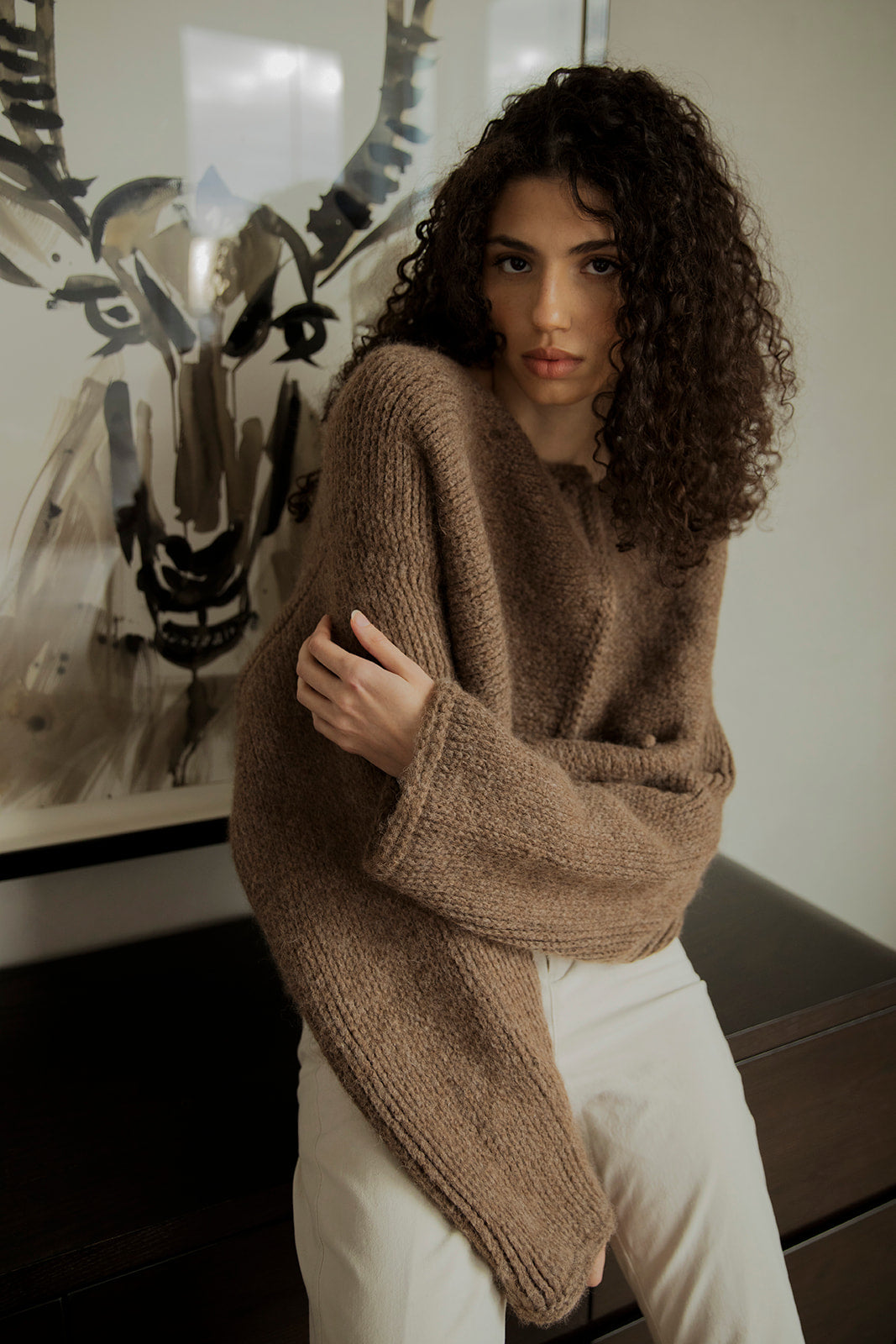 Model wearing the Momo Handknit Cardigan in Saddle, standing against a deer painting. The warm tones of saddle brown, blacks and creams of the painting complement the cardigan.