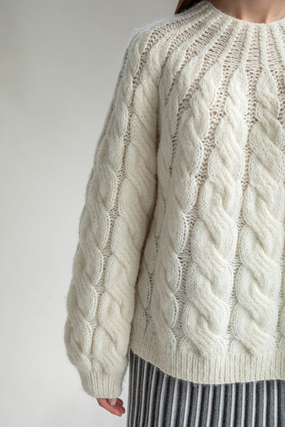 Close-up of the intricate cable-knit pattern on the Tomo Mix Cable Handknit. The soft texture of the ivory yarn is visible, showing the luxurious feel and craftsmanship of the hand-knit design by Peruvian artisans.