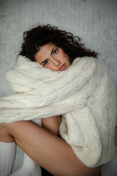 The cozy, oversized fit and soft, cable-knit texture are highlighted on the model as she nestles into the warmth of the Tomo Cable Mix Handknit, creating a relaxed moment that showcases its comfort.