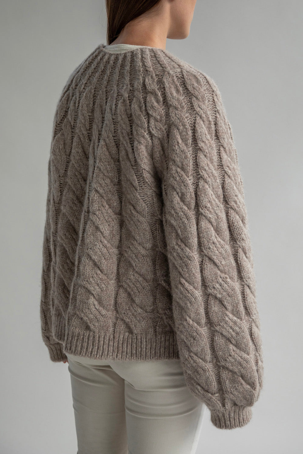 Side view of the Tomo Mix Cable Handknit made with a lightweight alpaca blend yarn. The pullover in Oat Heather shows a comfortable fit with ribbed detailing at the cuffs and hem. Available in one size.