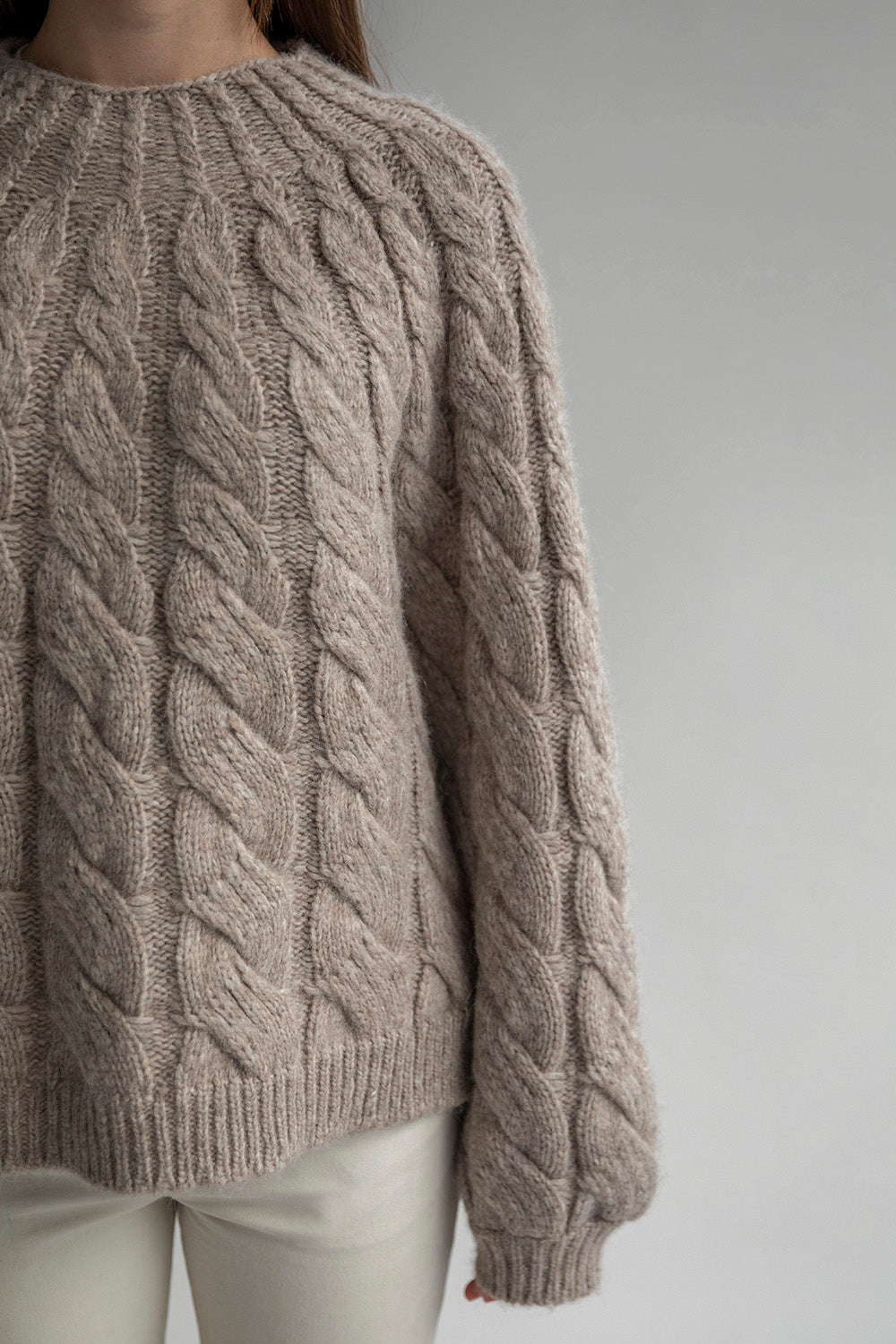 Close-up of the intricate cable-knit pattern on the Tomo Mix Cable Handknit. The soft texture of the oat heather yarn is visible, showing the luxurious feel and craftsmanship of the hand-knit design by Peruvian artisans.