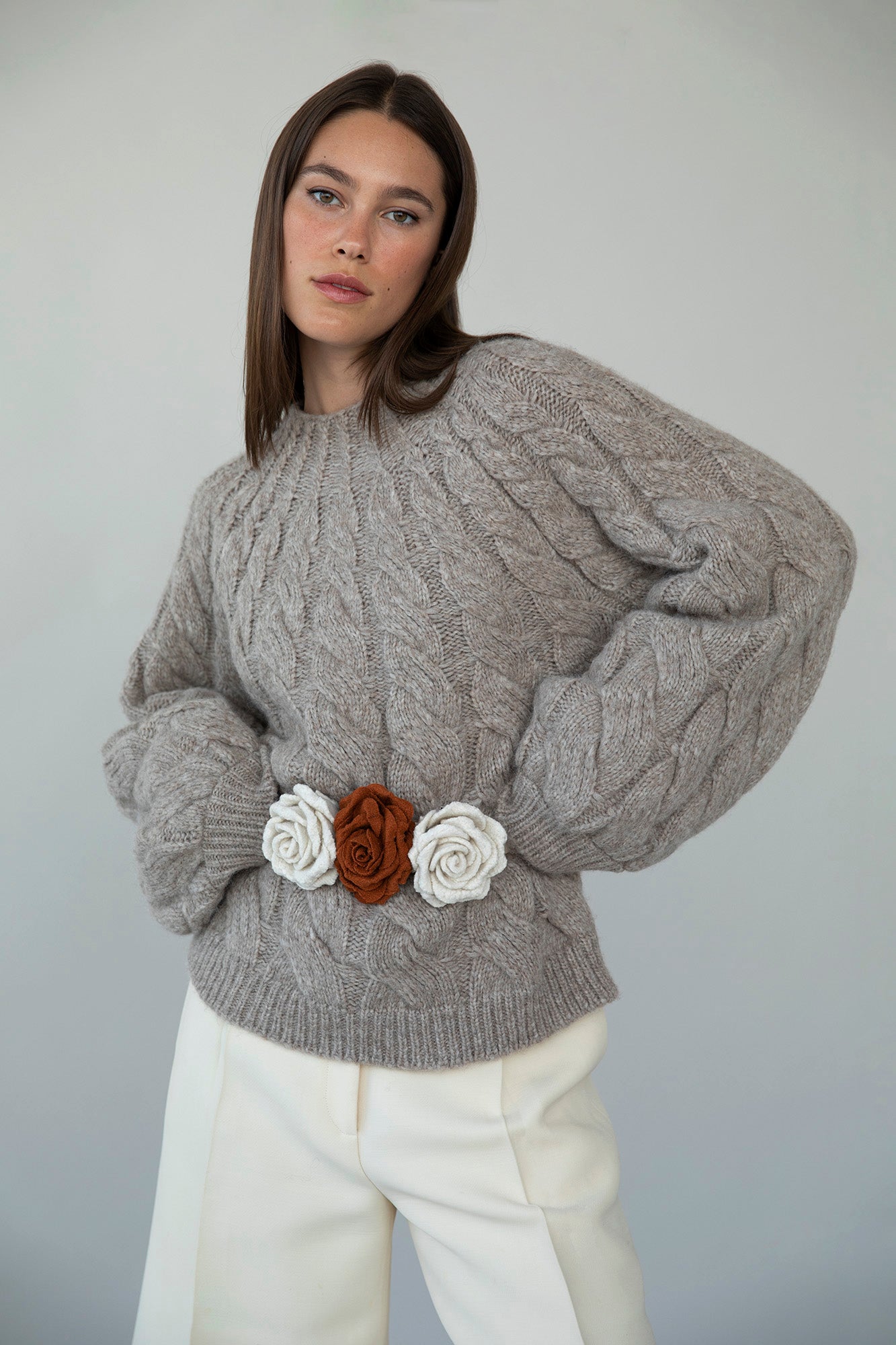 Model wearing the Tomo Mix Cable Handknit in Oat Heather, paired with three upcycled handmade rosettes styled as a belt around the sweater. The rosettes designed in collaboration with Ruari Studios x Sskein add a unique, artistic touch to the look.