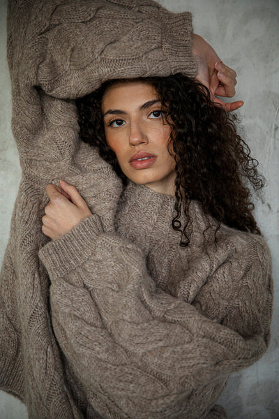 Model Bella in the Oat Heather color of the Tomo Mix Cable Handknit. Her arms are raised emphasizing the voluminous drama of the sleeves and textured warmth.